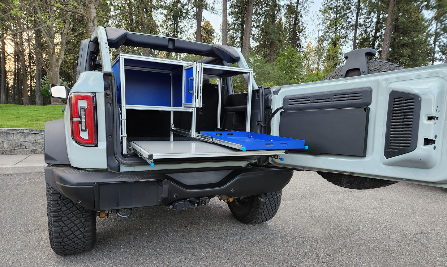 This is a photo of the Bronco Kitchen with the pantry door open, fridge and table slidouts are extended. Storage under the assebly is accessible and space is provided for recovery gear, food stock, and storage.